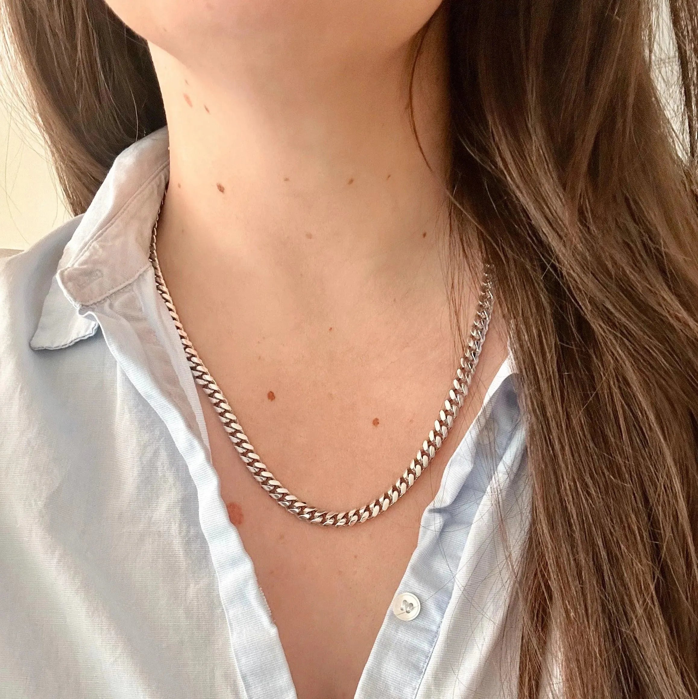 A person with long brown hair wears a Miami Cuban Link necklace from the Simply Silver Unisex collection. The light blue button-up shirt is open at the collar, emphasizing moles on their neck and chest—a touch of luxury in each detail.